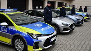 Pod ścianą w rzędzie stoją samochody policyjne. Dwa są oznakowane z napisem policja na drzwiach i masce samochodu. Pomiędzy autami stoją umundurowani policjanci.