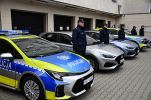 Pod ścianą w rzędzie stoją samochody policyjne. Dwa są oznakowane z napisem policja na drzwiach i masce samochodu. Pomiędzy autami stoją umundurowani policjanci.