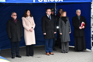 Pod niebieskim namiotem ogrodowym stoją w szeregu osoby ubrane z ciepłe zimowe kurtki i płaszcze. Na końcu szeregu stoi ksiądz ubrany w sutannę.
