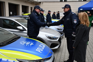 Pod ścianą w rzędzie stoją samochody policyjne. Dwa są oznakowane z napisem policja na drzwiach i masce samochodu. Pomiędzy autami stoją umundurowani policjanci. Jednemu policjantowi podaje dłoń inny umundurowany policjant, obok nich tyłem stoi kobieta ubrana w jasny zimowy płaszcz.