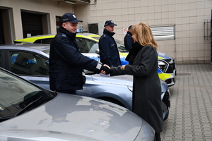 Pod ścianą w rzędzie stoją samochody policyjne. Dwa są oznakowane z napisem policja na drzwiach i masce samochodu. Pomiędzy autami stoją umundurowani policjanci. Jednemu policjantowi kobieta ubrana w zimowy płaszcz ściska dłoń, gratulując mu.