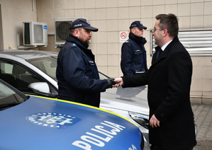 Pod ścianą w rzędzie stoją samochody policyjne. Dwa są oznakowane z napisem policja na drzwiach i masce samochodu. Pomiędzy autami stoją umundurowani policjanci. Jednemu policjantowi mężczyzna ubrany w zimowa kurtkę składa gratulacje ściskając dłoń.