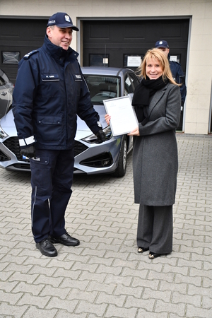 Na tle radiowozu policyjnego stoi wysoki umundurowany policjant, obok którego stoi kobieta w rozpuszczonych włosach i płaszczu zimowym. Kobieta w rękach trzyma ramkę z dyplomem w środku.