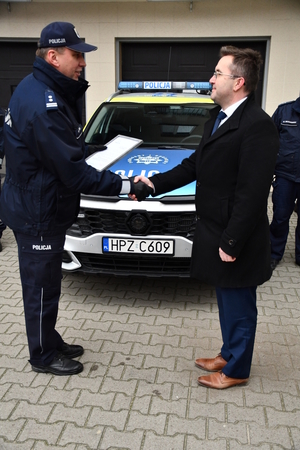 Na tle oznakowanego radiowozu policyjnego stoi wysoki umundurowany policjant, który w jednej ręce trzyma ramkę z dyplomem w środku, a drugą rękę podaje mężczyźnie ubranemu w zimową kurtkę.