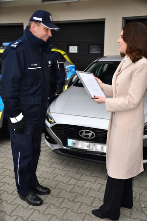 Na tle samochodu stoi wysoki umundurowany policjant który skierowany jest twarzą do stojącej na przeciwko niego kobiecie, która ubrana jest w zimowy płaszcz, ma rozpuszczone włosy zasłaniające twarz, a w ręku trzyma ramkę z dyplomem.