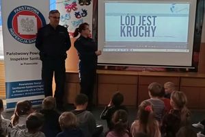 W pomieszczeniu stoi dwoje umundurowanych policjantów obok tablicy multimedialnej, na której jest napis: Lód jest kruchy. Z drugiej strony jest rozłożony baner z napisem Państwowa Inspekcja Sanitarna w Piasecznie oraz Powiatowa Stacja Sanitarno-Epidemiologiczna w Piasecznie z/s w Chylicach, ul. Dworska 7, 05-510 Konstancin -Jeziorna. Przed funkcjonariuszami siedzi grupa dzieci.