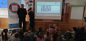 W pomieszczeniu stoi dwoje umundurowanych policjantów obok tablicy multimedialnej, na której jest napis: Lód jest kruchy. Z drugiej strony jest rozłożony baner z napisem Państwowa Inspekcja Sanitarna w Piasecznie oraz Powiatowa Stacja Sanitarno-Epidemiologiczna w Piasecznie z/s w Chylicach, ul. Dworska 7, 05-510 Konstancin -Jeziorna. Przed funkcjonariuszami siedzi grupa dzieci