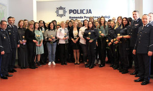 W pomieszczeniu stoi grupa osób. Prawie wszystkie to kobiety, które w ręku trzymają różę.Kobiety są ubrane zarówno w stroje cywilne jak i mundury policyjne. Wśród kobiet jest też nieliczne grono mężczyzn ubranych w większości w mundury policyjne.