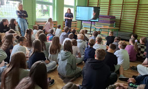 Umundurowana policjantka stoi pod ścianą w klasie. Twarzą zwrócona jest do dużej grupy uczniów, którzy siedzą na podłodze. Obok policjantki na ścianie wisi duży telewizor.