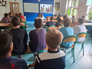 W szkolnej klasie siedzą uczniowie na krzesełkach. Zwróceni przodem do tablicy i wyświetlacza multimedialnego, który jest na ścianie.