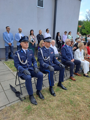 Dwaj umundurowani policjanci siedzą na krzesełkach, które stoją na trawie. Wokół stoją inni ludzie, niektórzy siedzą też na krzesełkach.