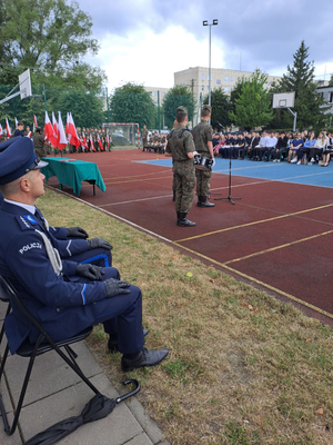 Umundurowany policjant siedzi na krzesełku. Przed nim jest boisko do gry w kosza, na którym jest mikrofon, przed nim stoi uczeń w zielonym mundurze i czyta tekst. w oddali stoi grupa uczni wraz z nauczycielami.