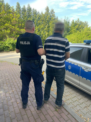 zatrzymany mężczyzna wraz z policjantem, obok stojący radiowóz policyjny