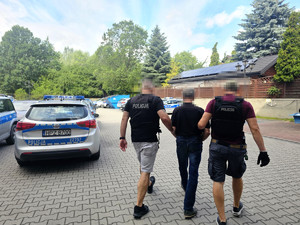 zatrzymany mężczyzna w asyście nieumundurowanych policjantów, zdjęcie wykonane przed jednostką, na parkingu