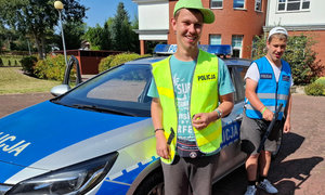 Dwóch nastolatków stoi obok oznakowanego radiowozu. Każdy z nastolatków ma na sobie kamizelkę z napisem policja. Jedna jest w kolorze żółtym, a druga w kolorze niebieskim. Obaj chłopcy maja na sobie bluzki z krótkim rękawem i czapkę z daszkiem.