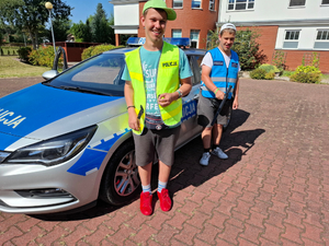 Dwóch nastolatków stoi obok oznakowanego radiowozu. Każdy z nastolatków ma na sobie kamizelkę z napisem policja. Jedna jest w kolorze żółtym, a druga w kolorze niebieskim. Obaj chłopcy maja na sobie bluzki z krótkim rękawem i czapkę z daszkiem.