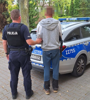 Umundurowany policjant trzyma pod rękę zatrzymanego mężczyznę. Obaj mężczyźni stoją tyłem ale przodem do oznakowanego radiowozu.