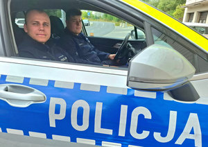 Dwóch umundurowanych policjantów siedzi w oznakowanym radiowozie z napisem policja.