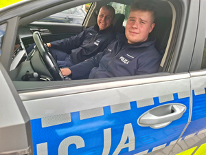Dwóch umundurowanych policjantów siedzi w oznakowanym radiowozie z napisem policja.