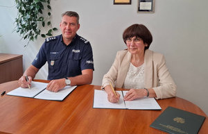 Umundurowany policjant siedzi razem przy stole wraz z kobietą ubrana w jasną bluzkę oraz sweter. Oboje siedzą obok siebie. Na stole przed nimi są rozłożone dokumenty, które podpisują.