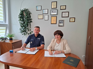 Umundurowany policjant siedzi razem przy stole wraz z kobietą ubrana w jasną bluzkę oraz sweter. Oboje siedzą obok siebie. Na stole przed nimi są rozłożone dokumenty, które podpisują.