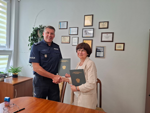 Umundurowany policjant gratuluje kobiecie uściskiem dłoni. Oboje stoją i trzymają w rękach teczki. Stoją przy stole, a za nimi jest ścianka, na której wiszą obrazki z podziękowaniami.Kobieta jest ubrana w jasna bluzkę i sweter.