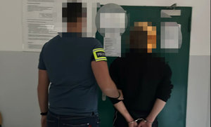 zatrzymany mężczyzna z kajdankami założonymi na ręce trzymane z tyłu w asyście policjanta, w tle wejście do pomieszczenia dla osób zatrzymanych