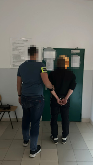 zatrzymany mężczyzna z kajdankami założonymi na ręce trzymane z tyłu w asyście policjanta, w tle wejście do pomieszczenia dla osób zatrzymanych