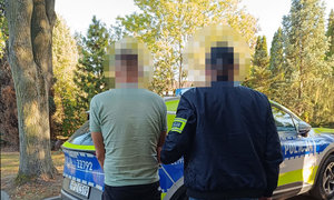 zatrzymany mężczyzna wraz z policjantem, w tle policyjny oznakowany radiowóz