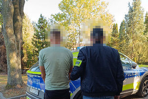 zatrzymany mężczyzna wraz z policjantem, w tle policyjny oznakowany radiowóz