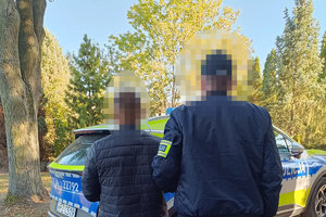 zatrzymany mężczyzna wraz z policjantem, w tle policyjny oznakowany radiowóz