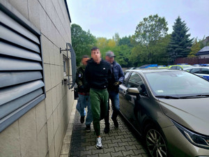 zatrzymany mężczyzna w asyście policjantów wydziału kryminalnego, zdjęcie wykonane na policyjnym parkingu, w tle radiowozy