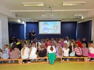 policjantki stojące przed dziećmi, które siedzą na ławkach i oglądają bajkę o bezpieczeństwie