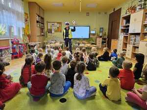 spotkanie w przedszkolu, dzieci siedzą na dywanie, policjant prowadzi spotkanie stojąc przed nimi