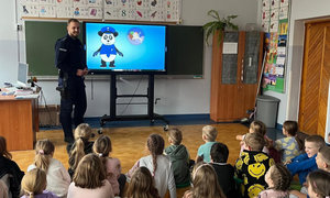 policjant stojący przed dziećmi, które siedzą na dywanie. w tle widać bajkę o bezpieczeństwie