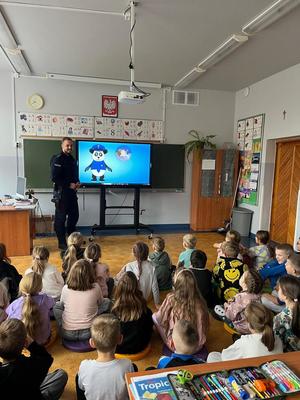 policjant stojący przed dziećmi, które siedzą na dywanie. w tle widać bajkę o bezpieczeństwie