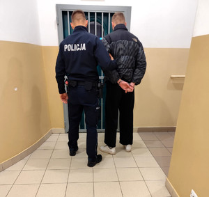 zatrzymany mężczyzna w asyście umundurowanego policjanta, na tle wejścia do pomieszczenia dla osób zatrzymanych