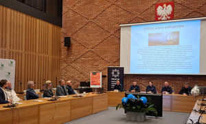 zdjęcie z debaty społecznej, sala konferencyjna. Przy stolikach siedzą przedstawiciele mieszkańców, urzędu, a także policjanci