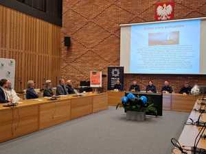 zdjęcie z debaty społecznej, sala konferencyjna. Przy stolikach siedzą przedstawiciele mieszkańców, urzędu, a także policjanci