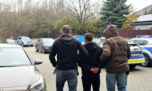 zatrzymany mężczyzna ma założone kajdanki na ręce trzymane z tyłu, idzie w asyście dwóch nieumundurowanych policjantów, zdjęcie wykonane na dziedzińcu wewnętrznym komendy, w tle policyjne oznakowane i nieoznakowane radiowozy