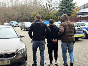 zatrzymany mężczyzna ma założone kajdanki na ręce trzymane z tyłu, idzie w asyście dwóch nieumundurowanych policjantów, zdjęcie wykonane na dziedzińcu wewnętrznym komendy, w tle policyjne oznakowane i nieoznakowane radiowozy