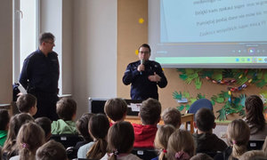 policjantka mówiąc do dzieci przez mikrofon, obok niej stoi drugi umundurowany policjant, uczniowie siedzą na krzesłach, w tle widać prezentację multimedialną
