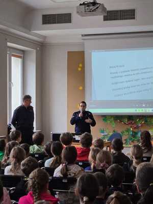 policjantka mówiąc do dzieci przez mikrofon, obok niej stoi drugi umundurowany policjant, uczniowie siedzą na krzesłach, w tle widać prezentację multimedialną