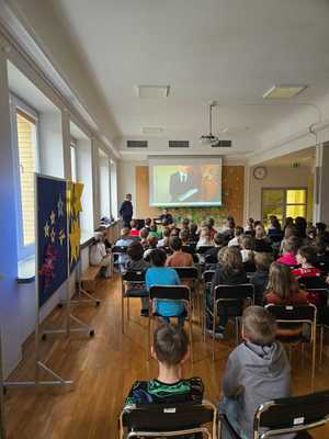 na zdjęciu widać uczniów siedzących na krzesłach, przed nimi stoją funkcjonariusze