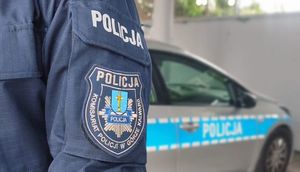 policjant z logiem Komisariatu Policji w Górze Kalwarii na mundurze, w tle radiowóz policyjny