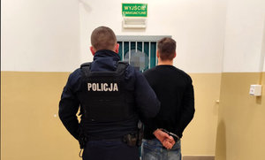 zatrzymany mężczyzna w asyście policjanta, w tle pomieszczenie dla osób zatrzymanych