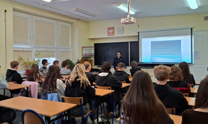 policjantka w trakcie spotkania z młodzieżą, zdjęcie wykonane w sali lekcyjnej. Policjantka stoi przy tablicy, uczniowie siedzą przy ławkach szkolnych, trwa prelekcja