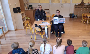 Na dywanie siedzą przedszkolne dzieci i patrzą na dwoje umundurowanych policjantów, którzy siedzą naprzeciwko nich na małych krzesełkach. Policjantka trzyma w reku dwie kartki. Na jednej jest znak przejścia dla pieszych a na drugiej jest całe przejście dla pieszych bez sygnałów świetlnych. Obok policjantki przy stoliku siedzi umundurowany policjant.