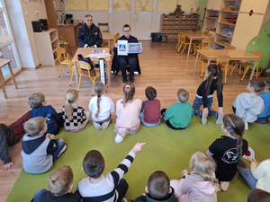 Na dywanie siedzą przedszkolne dzieci i patrzą na dwoje umundurowanych policjantów, którzy siedzą naprzeciwko nich na małych krzesełkach. Policjantka trzyma w reku dwie kartki. Na jednej jest znak przejścia dla pieszych a na drugiej jest całe przejście dla pieszych bez sygnałów świetlnych. Obok policjantki przy stoliku siedzi umundurowany policjant.