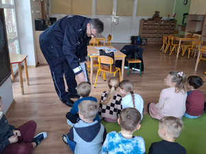 Na dywanie siedzą dzieci przedszkole. Wśród nich jest umundurowany policjant, który w ręku ma tubę. Do tej tuby zaglądają pojedynczo dzieci.